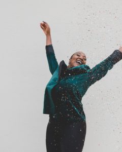 A woman in a teal blazer throws confetti while smiling at the sky and jumping with a white background.