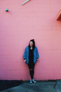 A woman in all black and a jean jacket leans back, laughing against a pink wall